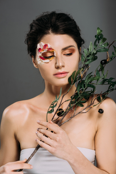 beautiful woman with petals on face holding green branch isolated on grey - Φωτογραφία, εικόνα
