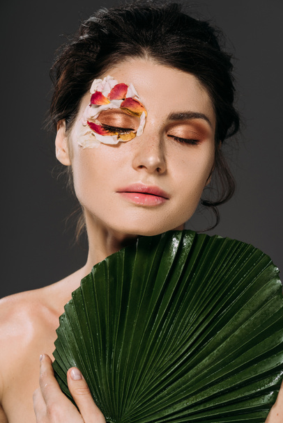 tender young woman with closed eyes and floral petals on face holding tropical leaf isolated on grey - Фото, зображення