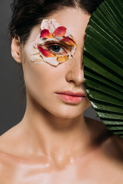 attractive girl with floral petals around eye holding leaf isolated on grey - Foto, afbeelding
