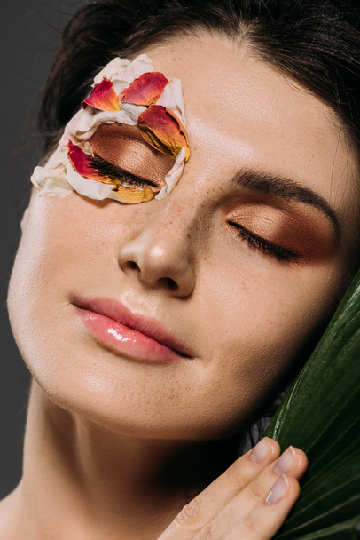 beautiful young woman with closed eyes with makeup and floral petals on face holding leaf isolated on grey - Foto, afbeelding