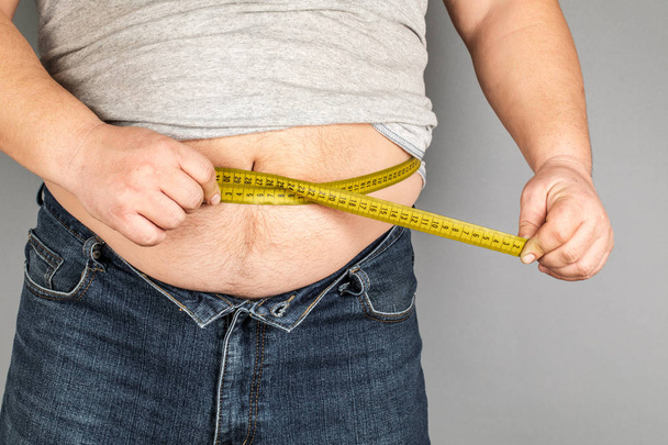 Muž měří jeho tlusté břicho měřicí páskou. na šedém pozadí - Fotografie, Obrázek