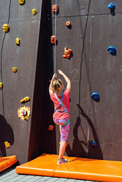 das verängstigte Mädchen ist blond, will mit Hilfe eines Sicherheitsseils auf einen künstlichen Felsen klettern, ist mit Klettern beschäftigt. - Foto, Bild