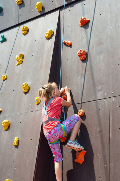 The frightened girl is blonde, wants to climb up on an artificial rock with the help of a safety rope, is engaged in rock-climbing. - Foto, Imagen