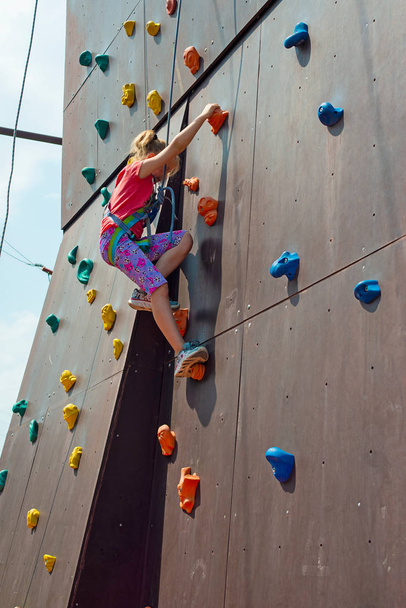 The girl is blonde, climbs to the top on an artificial rock with the help of a safety rope, is engaged in rock-climbing - Foto, imagen