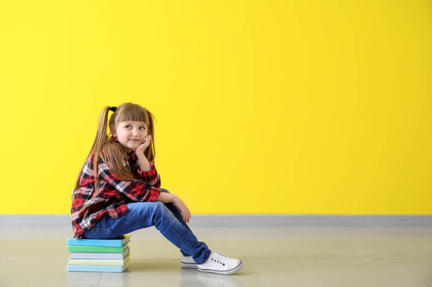 Χαριτωμένο μικρό κορίτσι που κάθεται στη στοίβα των βιβλίων κατά χρώμα τοίχου - Φωτογραφία, εικόνα