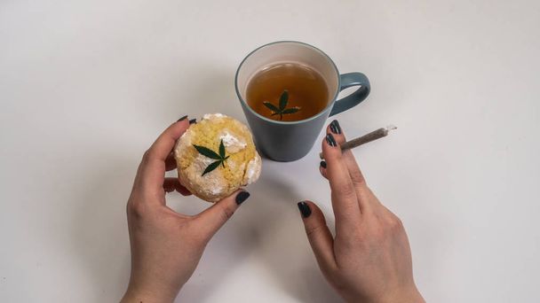 Pastelitos caseros (pastel) con marihuana medicinal y una tapa de té con hojas de marihuana sobre fondo blanco. Articulación de cannabis y pastel en mano de mujer
 - Foto, imagen