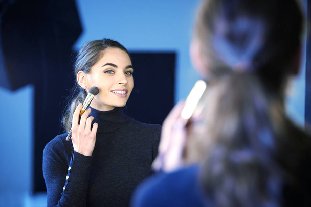 Portrait of gorgeous young woman applying her makeup while sitting at mirror in the photostudio. - Foto, Bild