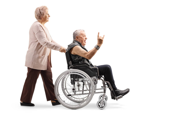 Perfil de comprimento total de uma mulher idosa empurrando um homem maduro fazendo um sinal de mão de rock and roll em uma cadeira de rodas isolada em fundo branco
 - Foto, Imagem