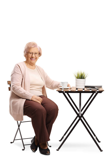 Full length portrait of a senior woman sitting at a table with a cup of coffee isolated on white background - Φωτογραφία, εικόνα