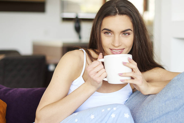 Close-up portrait shot of young woman wearing pajamas and drinking her morning tea while relaxing on sofa at home.  - Fotoğraf, Görsel