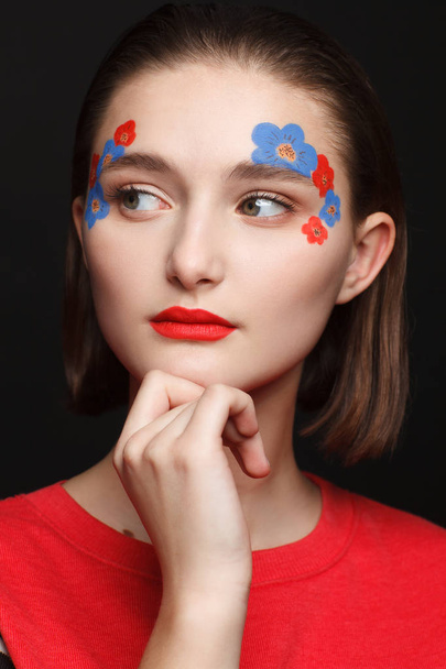 Photo of a young girl with makeup in the form of flowers in red and blue. Black background - Foto, Imagen