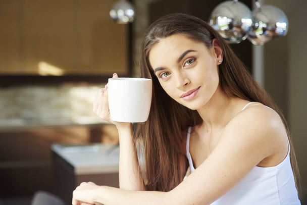 Close-up portrait shot of young woman wearing pajamas and drinking her morning tea while relaxing at home.  - Foto, afbeelding