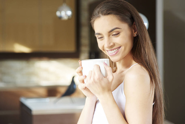 Portrait shot of happy woman wearing pajamas and drinking her morning tea while daydreaming at home. - Foto, Imagem