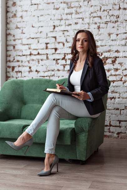 portrait of woman business coach sitting on the sofa shelf holding a notebook looking straight in lifestyle suit - Photo, Image