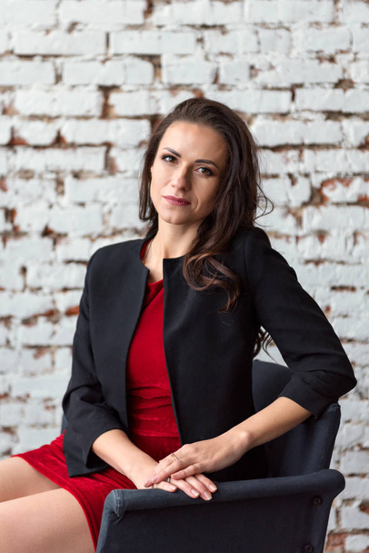 Portrait of a middle-aged brunette business lady in a short red dress and black jacket sitting on a chair with a white wall on the background. - Foto, Imagen