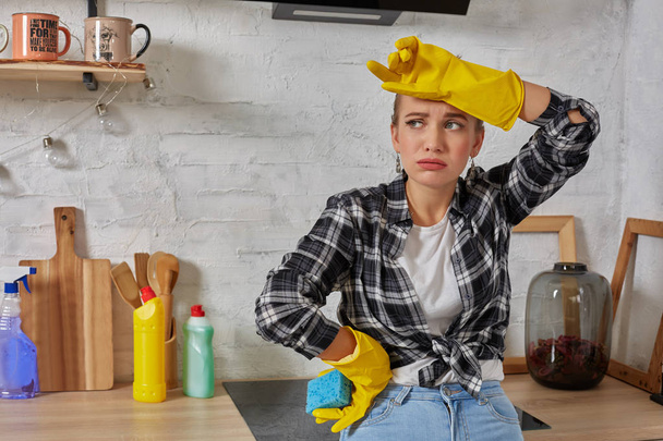 Blondine vrouw in beschermende handschoenen met vodden schoonmaken elektrisch fornuis thuis keuken. Meisje wassen zwart glanzend oppervlak van keukenblad. Begrip huishoudelijk werk. - Foto, afbeelding