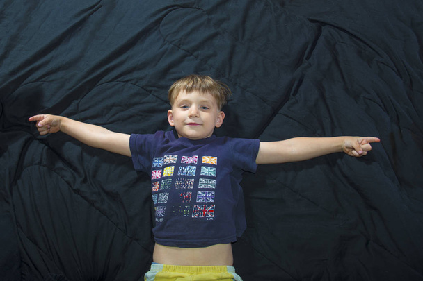 A little boy is lying on the couch and looks at the camera and raises his hands to the sides. - Photo, Image