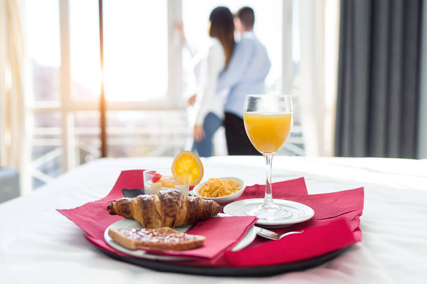 Close-up of continental breakfast in bedroom at hotel room. In the background; young couple looking cityscape through the window. - Φωτογραφία, εικόνα