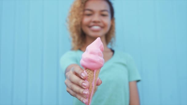 young afro girl eating pink melting ice cream at blue wall background - Imágenes, Vídeo