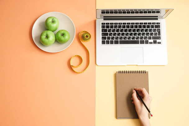 Female hand writing in notebook, laptop, apples and measuring tape on color background. Weight loss concept - Foto, immagini