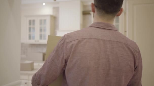 Bearded guy with glasses brings a cardboard box to the kitchen of his new home. Moving to a new apartment. - Séquence, vidéo