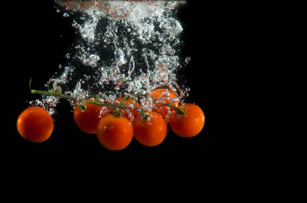 The branch of cherry tomatoes in water splash with bubbles on a black background. Photo through the water. - Photo, Image