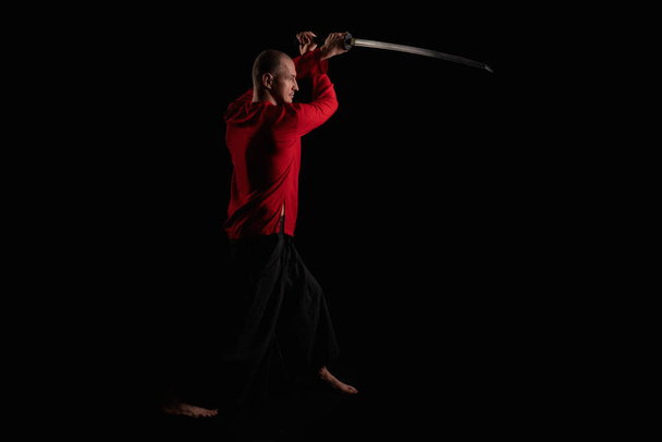 Man wearing red shirt doing exercises with a sword full-length profile shot in the dark room - Zdjęcie, obraz