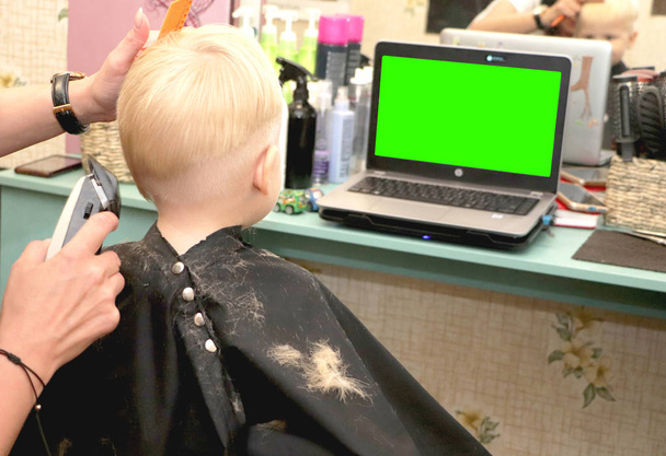 Un petit garçon coupe un coiffeur dans le salon. Le gamin regarde un dessin animé. Écran vert sur un ordinateur portable pour signature
 - Photo, image