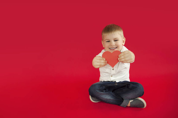 for holidays, lovers day and mothers day, baby boy sits on a red background with a red heart in his hands. The concept is to give gifts and love - Fotó, kép