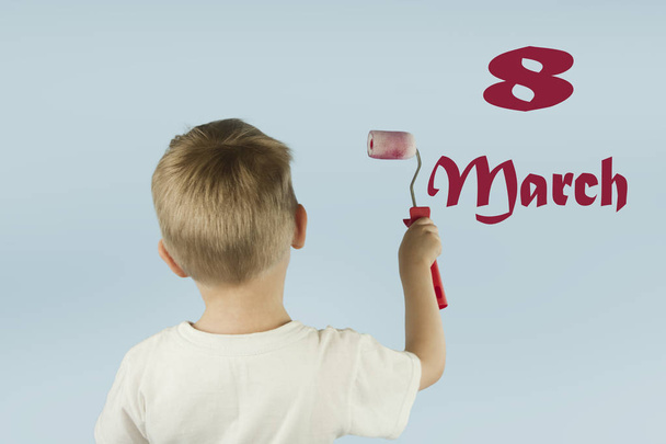 Hermoso niño rubio pinta la pared con un rodillo y escribe pintura en la pared sobre un fondo azul aislado. Concepto de descuentos y ventas
 - Foto, imagen