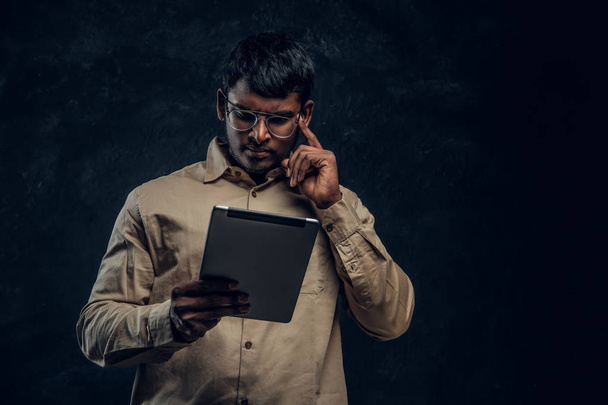 Pensive Indien mâle en lunettes et chemise à l'aide d'une tablette informatique en studio
 - Photo, image