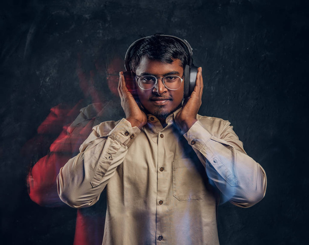 Bonito indiano cara está feliz ouvindo música em fones de ouvido sem fio de pé em um estúdio escuro
 - Foto, Imagem