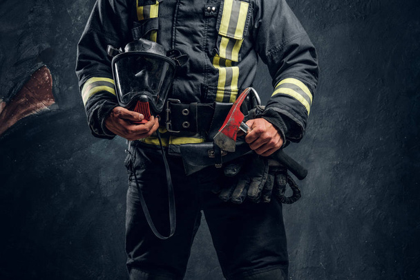 Cropped image of a male in uniform holding an oxygen mask and fire axe. Studio photo against a dark textured wall - Foto, afbeelding