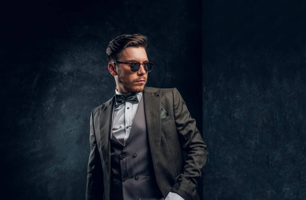 A stylish man in sunglasses dressed in an elegant suit posing with a hand in a pocket against a dark textured wall - Photo, Image