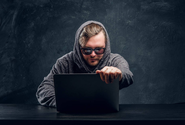 Ohjelmoija huppari ja aurinkolasit hakkerointi salasanoja kannettavan tietokoneen istuu pöydän pimeässä huoneessa
 - Valokuva, kuva