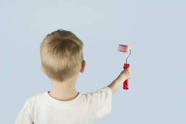 Beautiful blond child boy paints the wall with a roller and writes paint on the wall on an isolated blue background. Concept of discounts and sales - Foto, afbeelding