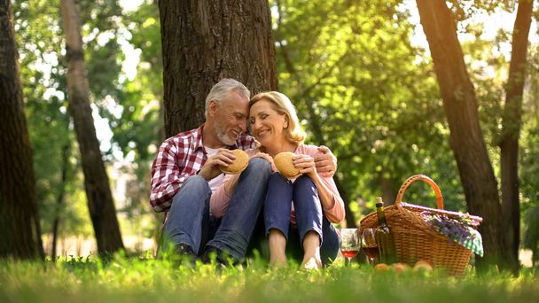 Cheerful old couple resting on grass and eating burgers, romantic date in park - Foto, Imagen