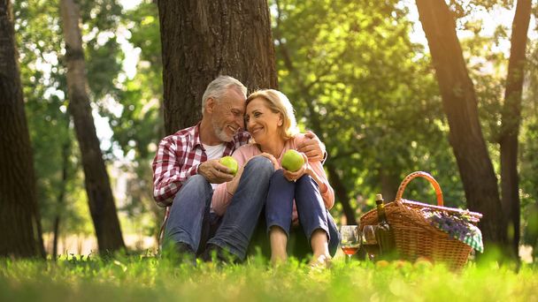Loving old couple hugging in park and eating green apples, picnic family weekend - Foto, Bild