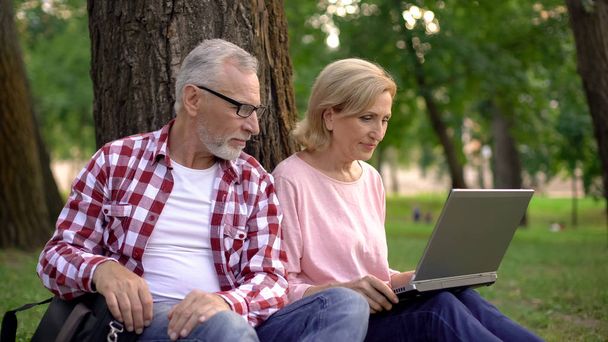 Male and female pensioners choosing hotel for vacation on laptop resting in park - Foto, Imagen