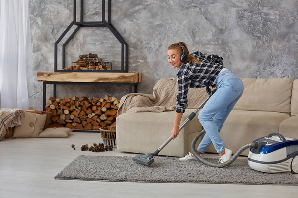 Full length portrait of cheerful young woman 20s listening to music via headphones and having fun with vacuum cleaner in the house. Busy, cleaning day. Home, housekeeping concept - Photo, Image