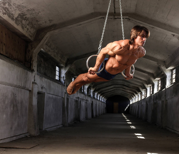 A professional sportsman with a beautiful muscular body trains on gymnastic rings in abandoned industrial building - Foto, immagini