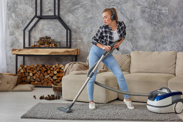 Full length portrait of cheerful young woman 20s listening to music via headphones and having fun with vacuum cleaner in the house. Busy, cleaning day. Home, housekeeping concept - Fotó, kép