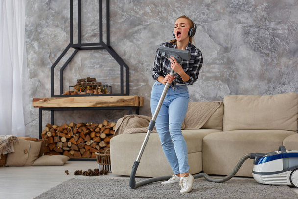 Cheerful young woman enjoying solo singing with vacuum cleaner while cleaning house. Busy, cleaning day. Home, housekeeping concept - 写真・画像