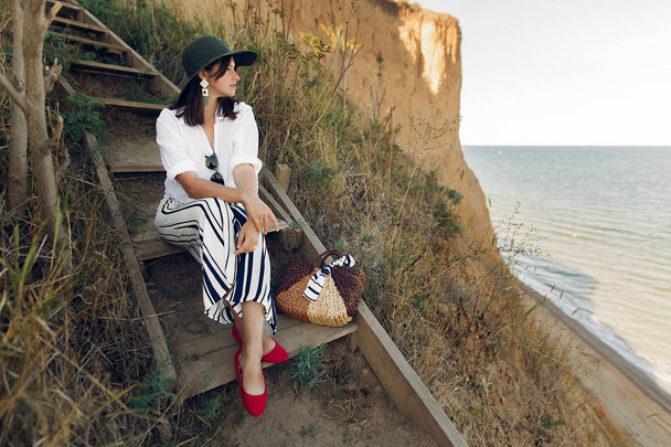 Κομψό κορίτσι στη μοντέρνα ματιά κάθεται σε ξύλινες σκάλες στον βράχο της παραλίας στη θάλασσα στο ηλιόλουστο φως. Χαρούμενος νεαρός boho γυναίκα με καπέλο με χαλαρωτικό αχυρένια τσάντα στην ξηρά στο ηλιοβασίλεμα. Καλοκαιρινές διακοπές. - Φωτογραφία, εικόνα