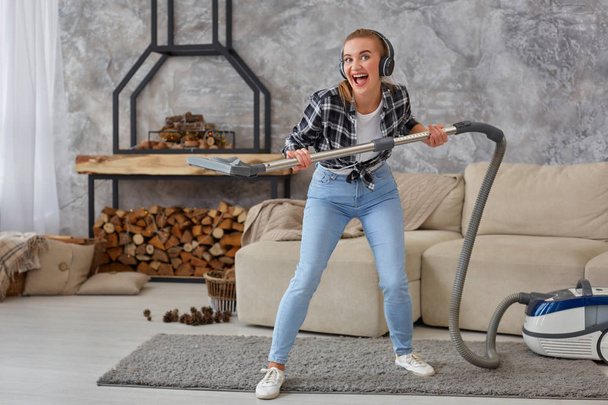 Full length portrait of cheerful young woman 20s listening to music via headphones and having fun with vacuum cleaner in the house. Busy, cleaning day. Home, housekeeping concept - Фото, изображение