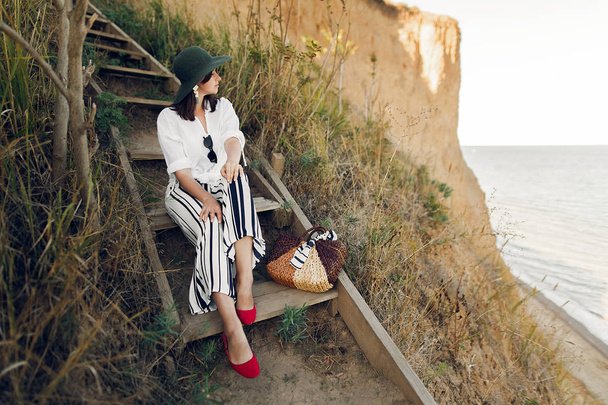 Стильная девушка модного вида, сидящая на деревянной лестнице на пляжной скале у моря в солнечном свете. Счастливая молодая женщина в шляпе с соломенным мешком, отдыхающая на берегу на закате. Летний отдых
. - Фото, изображение