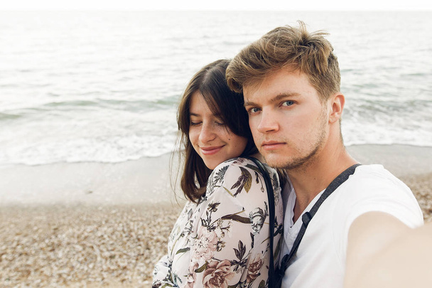 Stylish hipster couple taking selfie and embracing on beach in evening. Happy family relaxing on honeymoon at sea. Summer vacation. - Foto, immagini