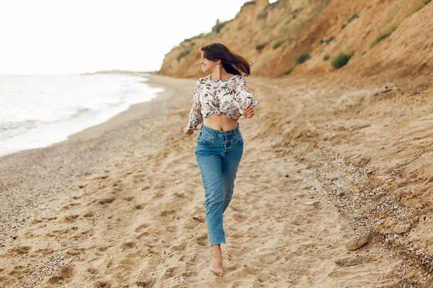 Счастливая молодая женщина бегает и веселится на солнечном пляже со скалой. Хипстерская стройная девушка расслабляется и улыбается на море. Летние каникулы. Скопируй пространство. Радостный момент
 - Фото, изображение