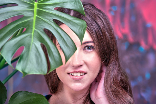 Close up portrait of a young brunette lady, wearing blue dress. The girl posing, looking at the camera. Green palm leaves surround her face - Foto, afbeelding