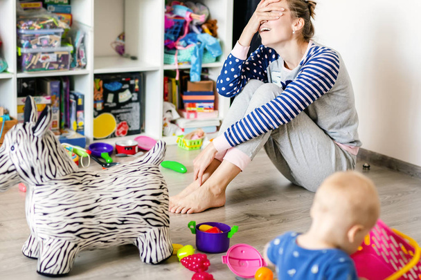 Tired of everyday household mother sitting on floor with hands on face. Kid playing in messy room. Scaterred toys and disorder. Happy parenting - Фото, изображение